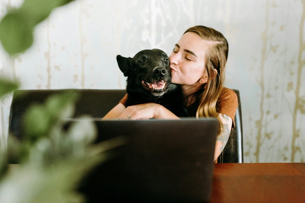A woman kissing her dog while sitting at a desk or table in front of her laptop. Both the dog and the woman are smiling.